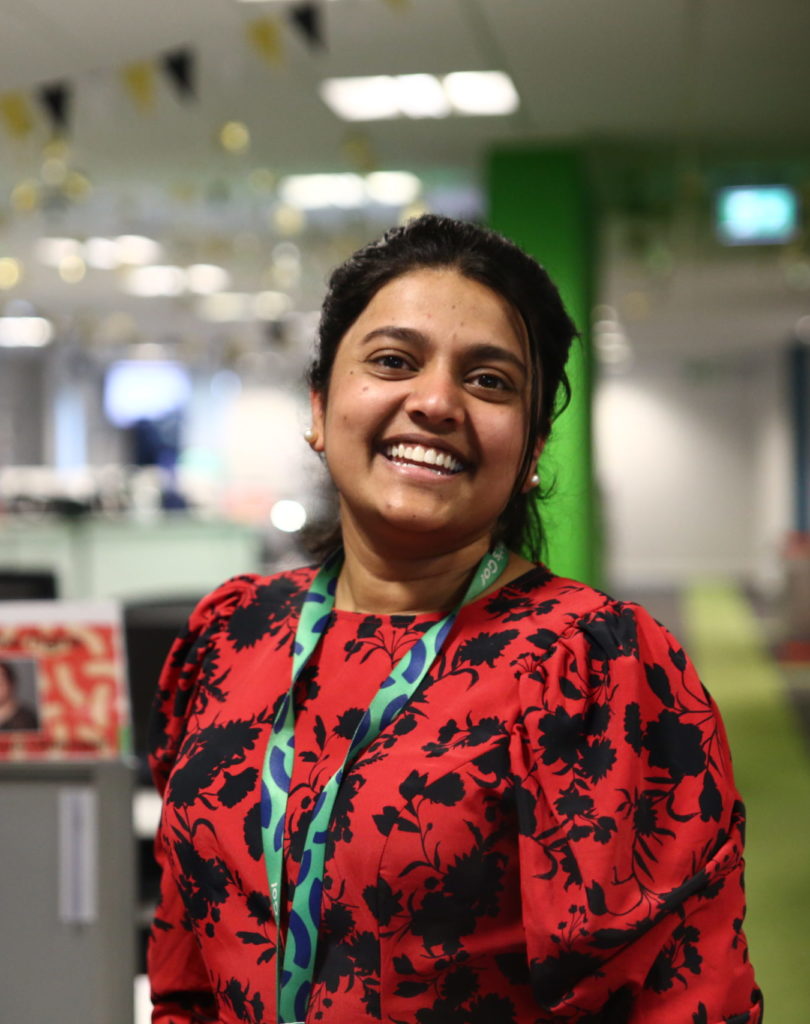 Sameea Bux smiling stood slightly side on in office space with desk blurred in the background