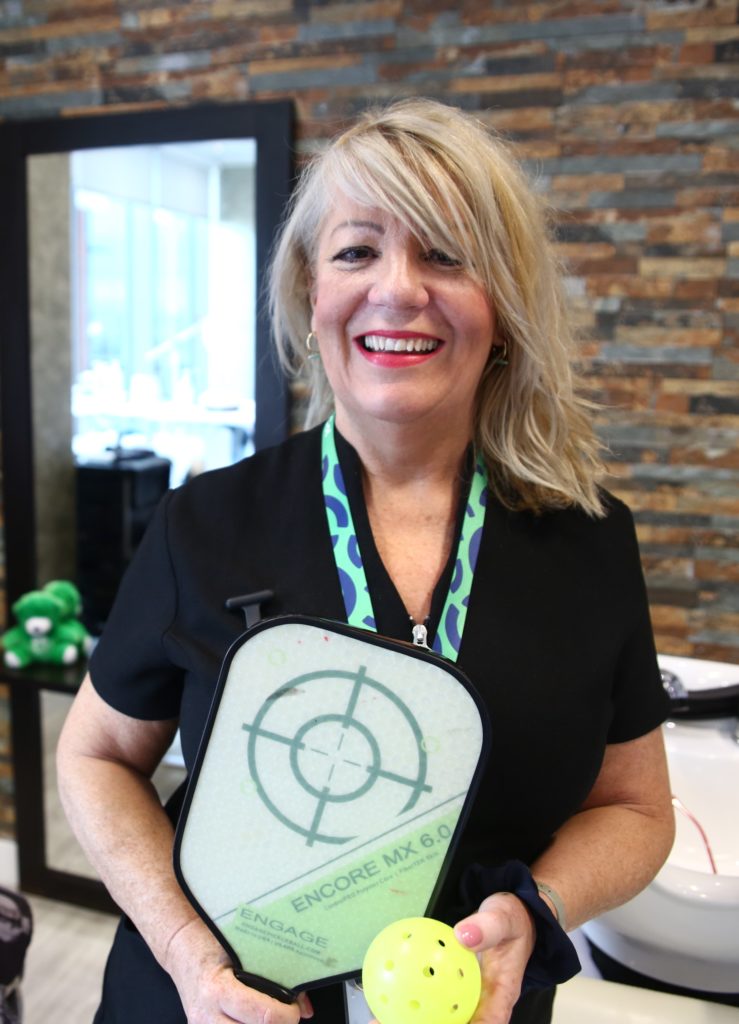 June in the spa smiling at the camera holding her Pickleball bat and ball with haircutting chair and mirror blurred in the background