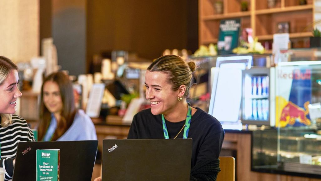 “Three people working on laptops and chatting at a table in a café-style co-working space.”
