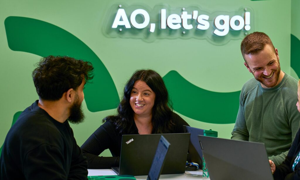 "Four people seated around a table with laptops, engaged in a collaborative discussion in front of a green wall displaying the text 'AO, let's go!'"