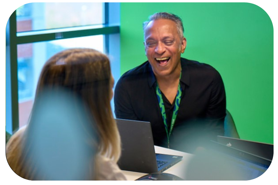 “Two colleagues working on laptops and talking in a bright office with green walls.”